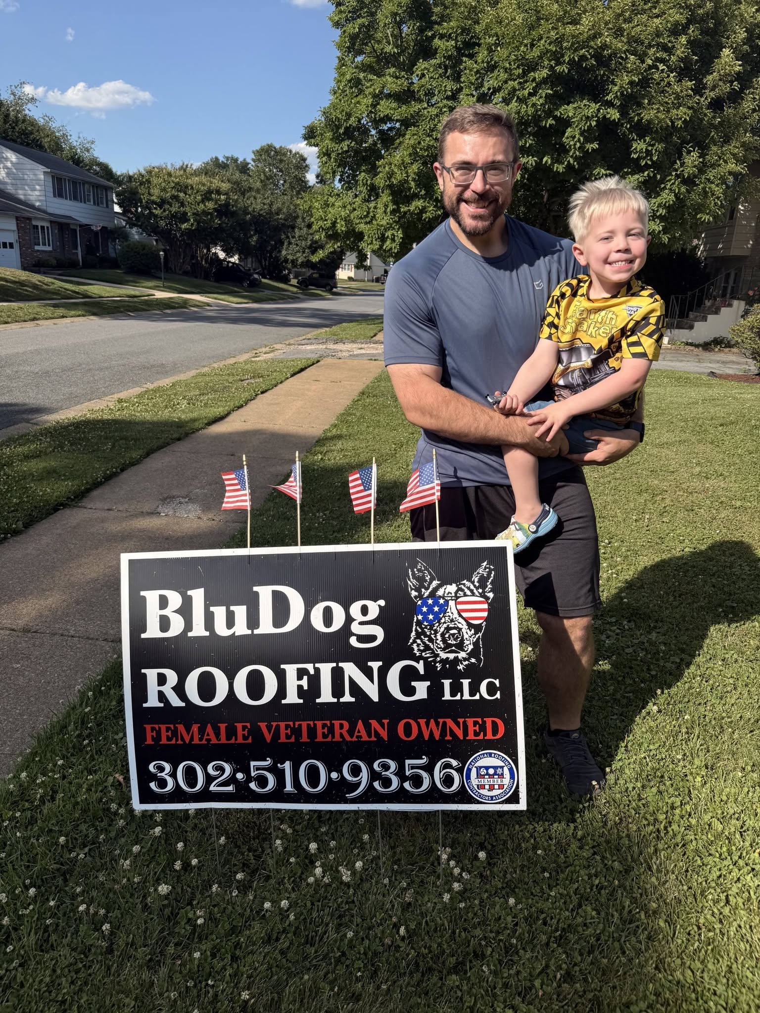 Father and child smiling beside a BluDog Roofing yard sign